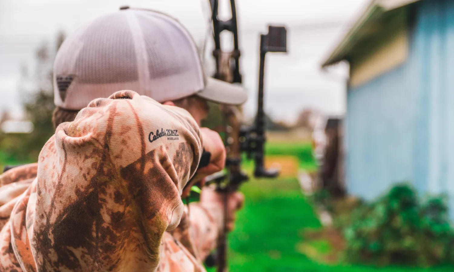 Archer in a camouflage hoodie and cap aiming a compound bow in a backyard, seen from behind