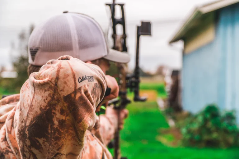 Archer in a camouflage hoodie and cap aiming a compound bow in a backyard, seen from behind