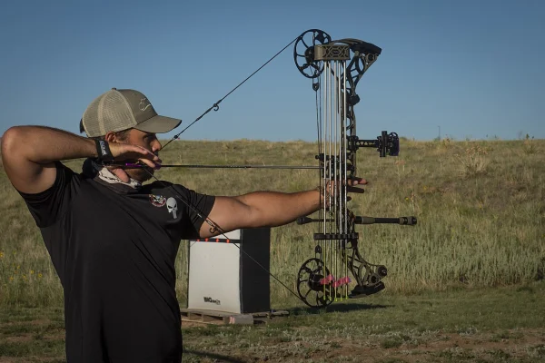 Archer in a cap drawing a compound bow fitted with a sight and stabilizer, using a release aid