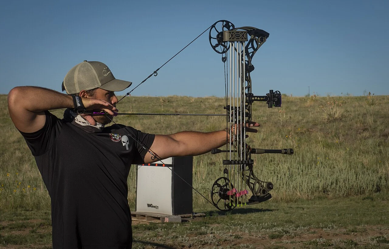 Archer in a cap drawing a compound bow fitted with a sight and stabilizer, using a release aid