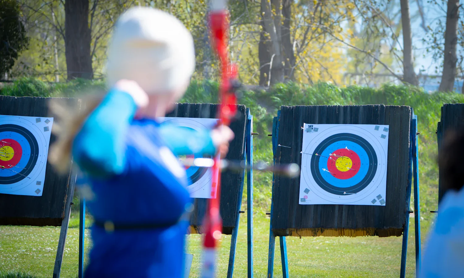 Archer in a blue jacket holding a recurve bow in front of target faces, seen from behind
