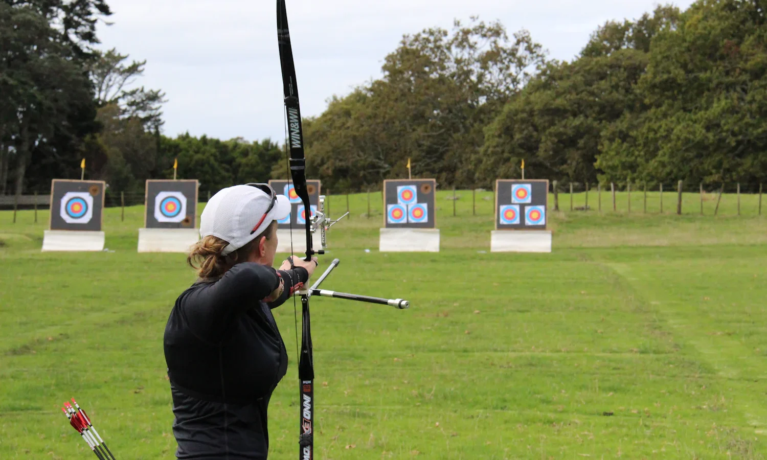 Archer with an Olympic-style recurve bow facing a row of targets on a grass range, seen from behind