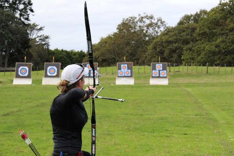 Archer with an Olympic-style recurve bow facing a row of targets on a grass range, seen from behind