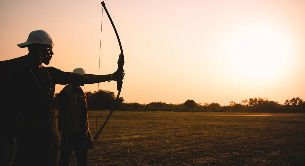 Silhouette of two archers, one at full draw with a recurve bow, against an orange sunset sky