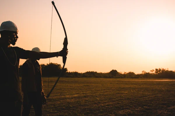Silhouette of two archers, one at full draw with a recurve bow, against an orange sunset sky