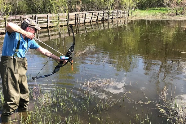 Bowfisher in waders drawing a bowfishing bow at the edge of a pond