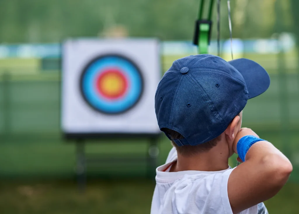 Child in a blue cap facing an archery target at an outdoor range, seen from behind
