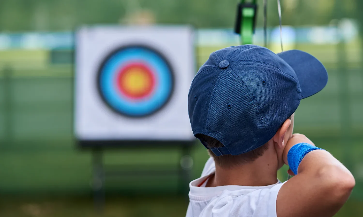 Child in a blue cap facing an archery target at an outdoor range, seen from behind