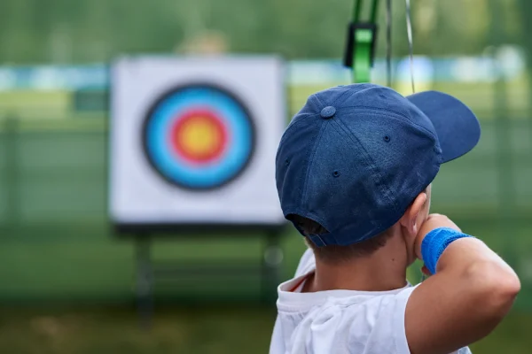 Child in a blue cap facing an archery target at an outdoor range, seen from behind