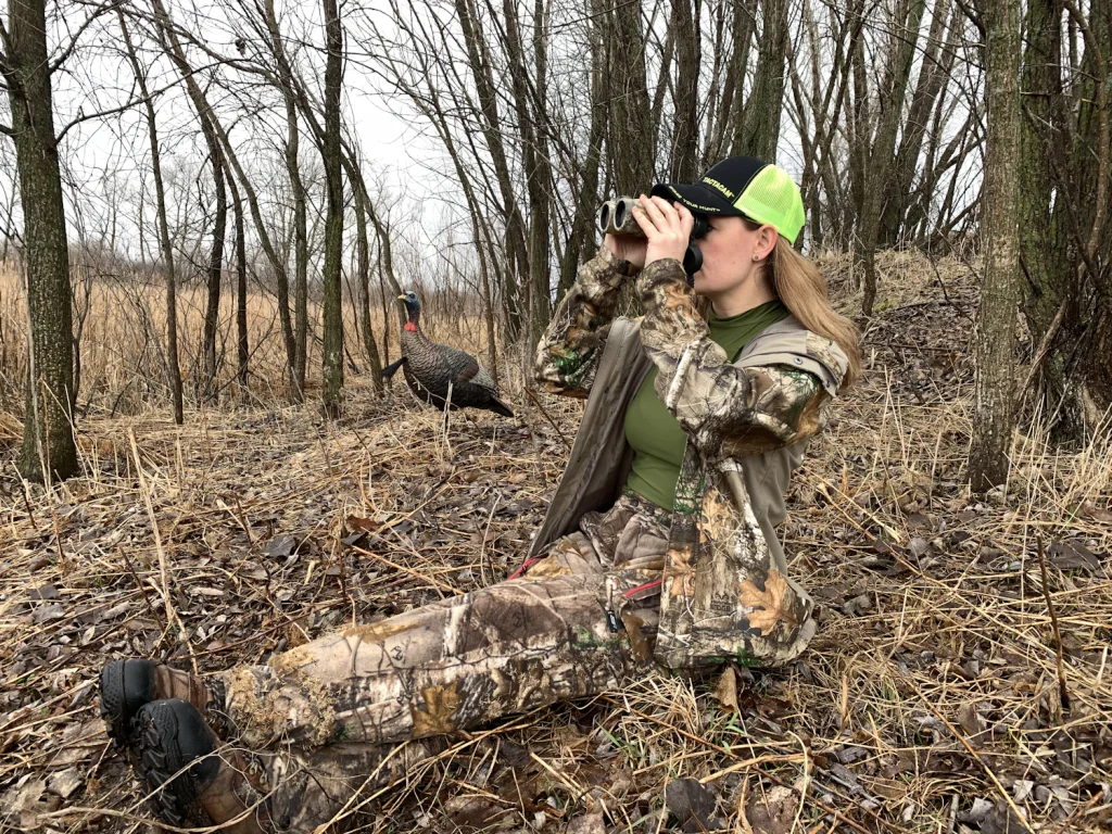 Hunter in camouflage sitting on the ground in bare woods and looking through binoculars