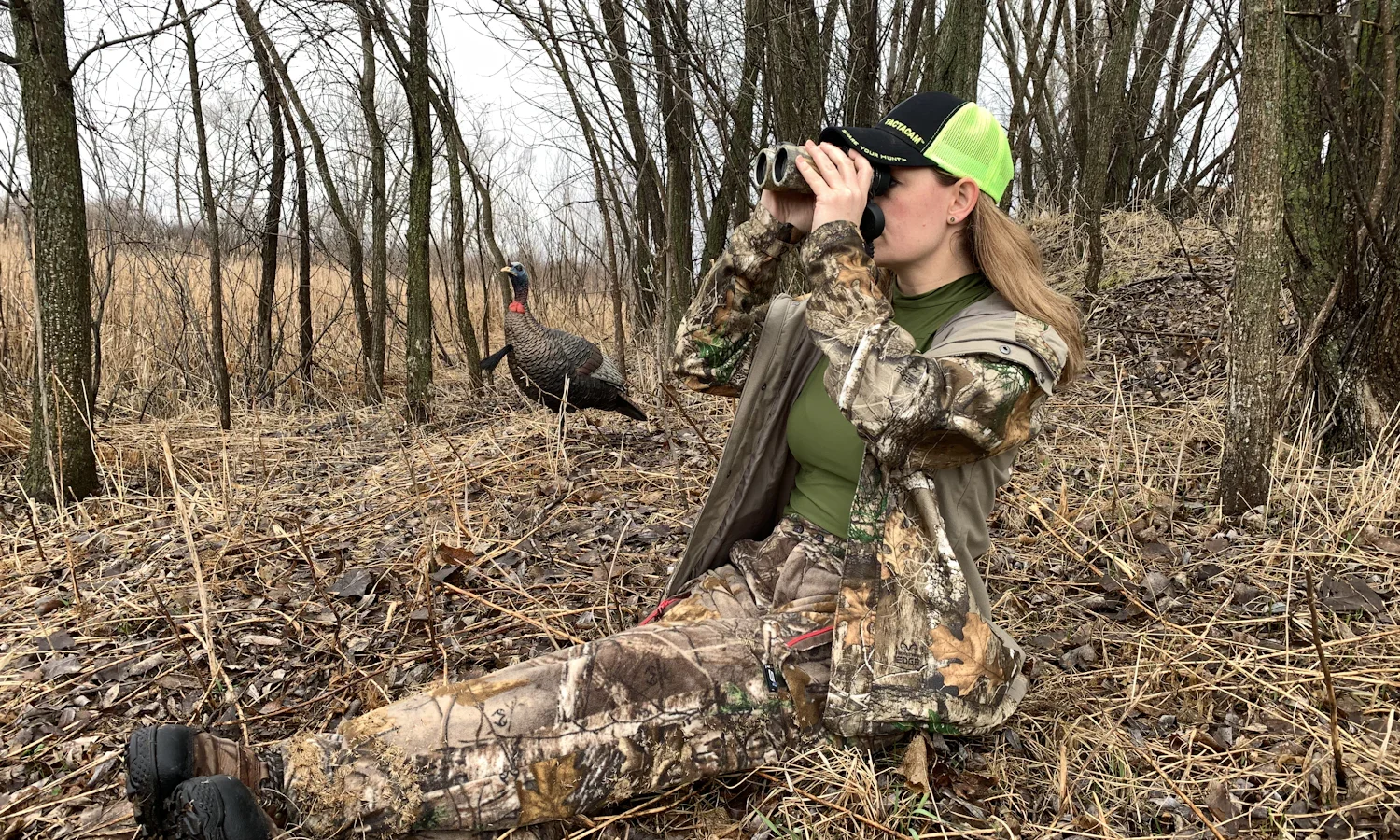 Hunter in camouflage sitting on the ground in bare woods and looking through binoculars