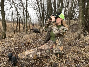 Hunter in camouflage sitting on the ground in bare woods and looking through binoculars