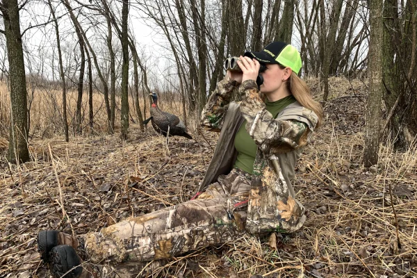 Hunter in camouflage sitting on the ground in bare woods and looking through binoculars