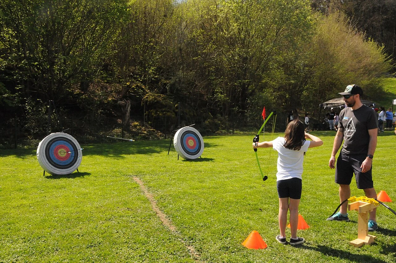 Instructor standing beside a child who is aiming a bow at targets on a grass range