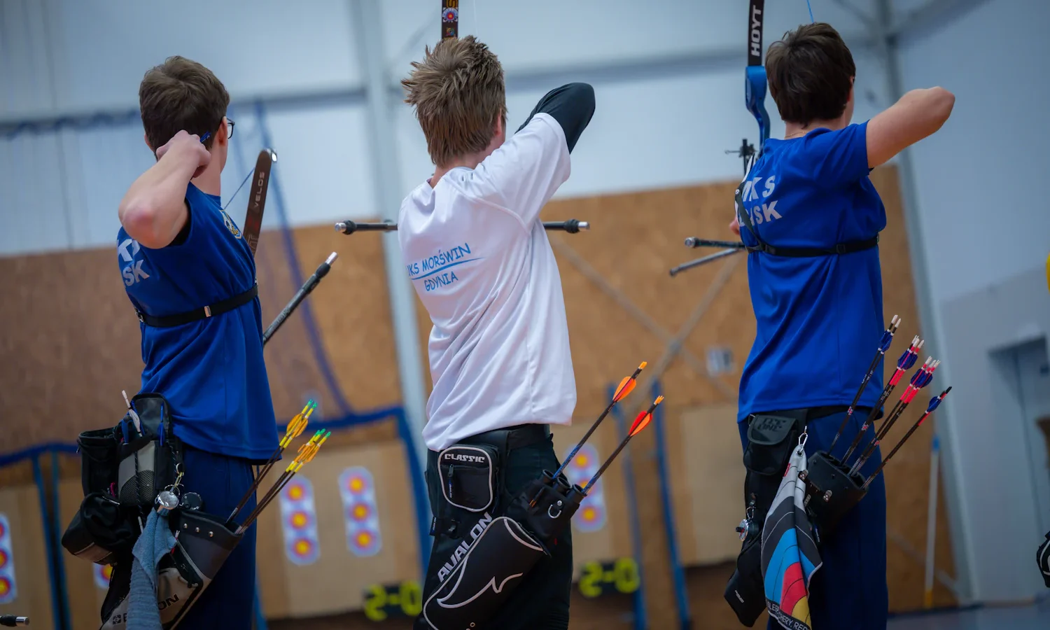 Three young archers shooting at an indoor range, seen from behind